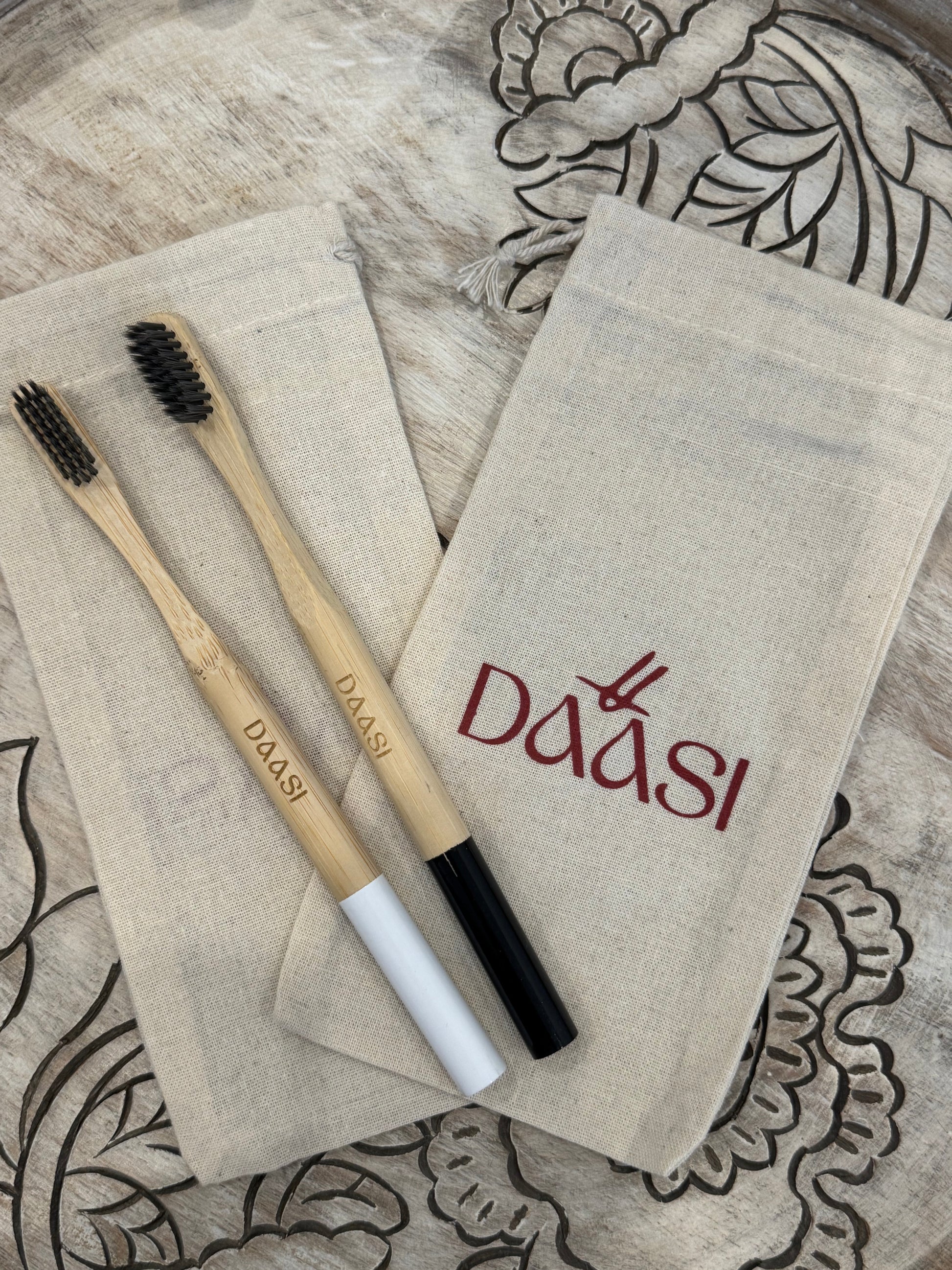 Two DAASI Dantya bamboo toothbrushes, one with a black handle and one with a white handle, laid across two DAASI branded canvas pouches on a carved wood tray